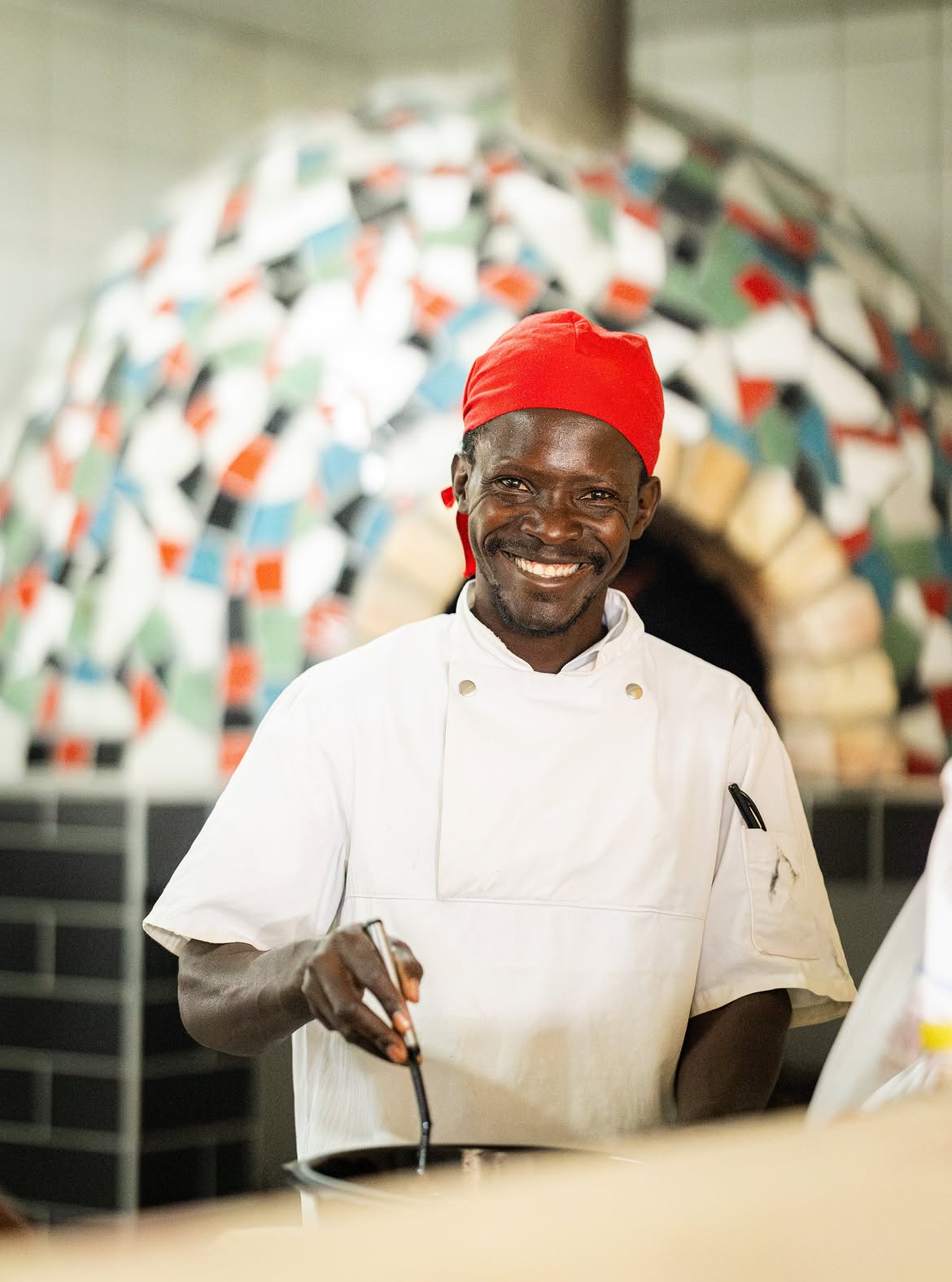 Chef in a red bandana smiling in front of the mosaic pizza oven