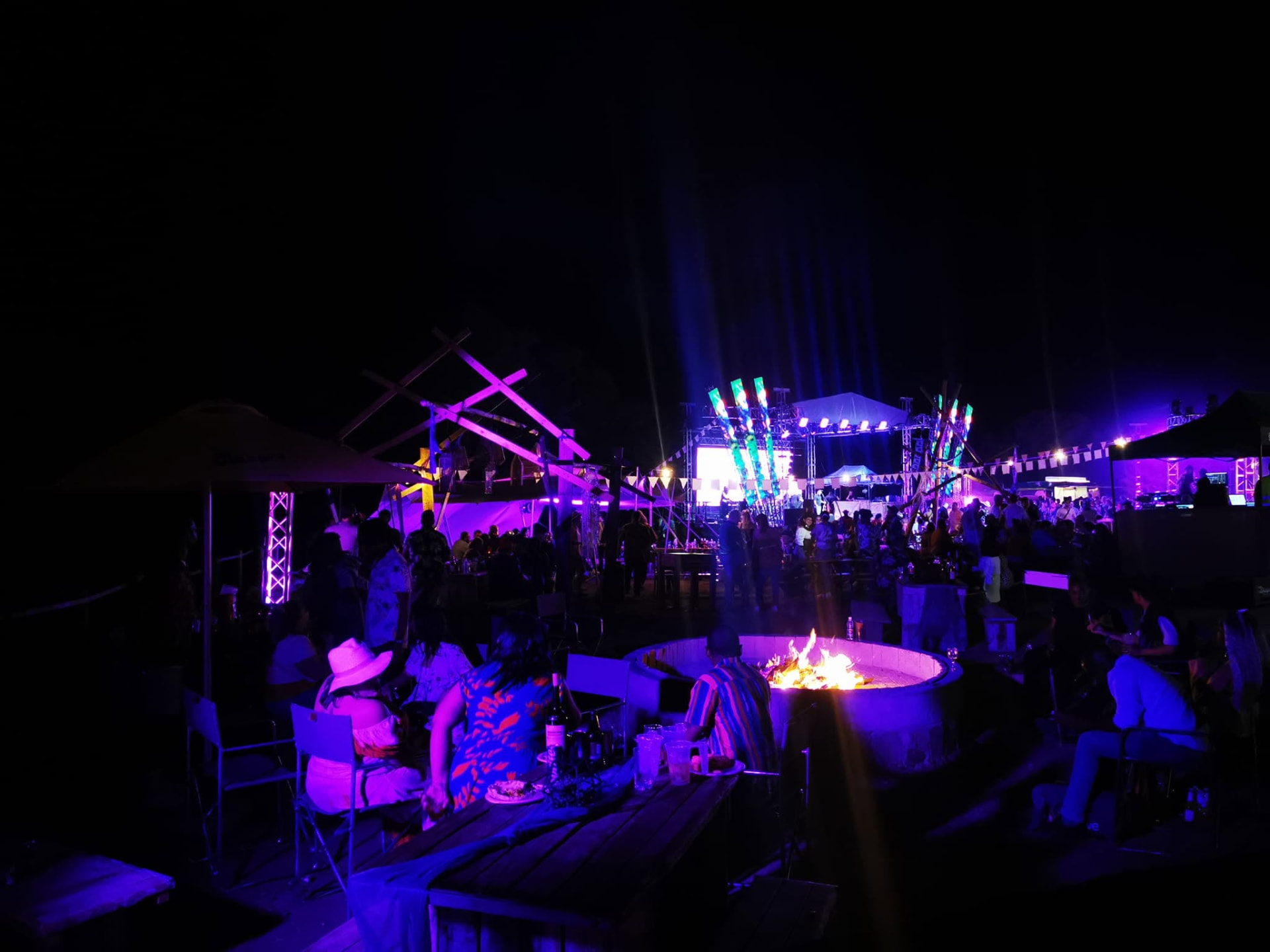 Guests around a lit fire pit at night with a stage and coloured lighting behind
