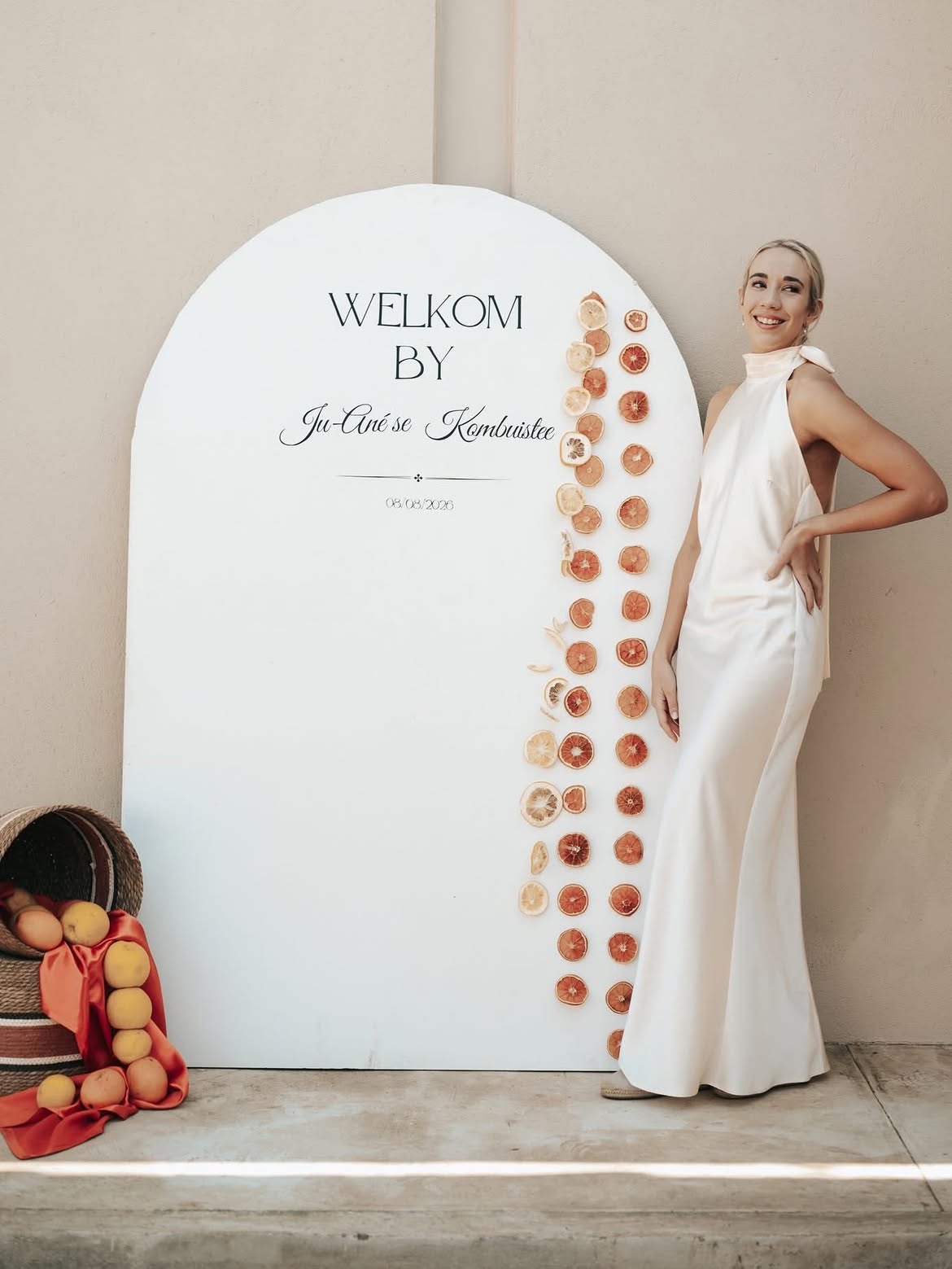 Bride in a satin dress beside an arched welcome sign trimmed with dried citrus