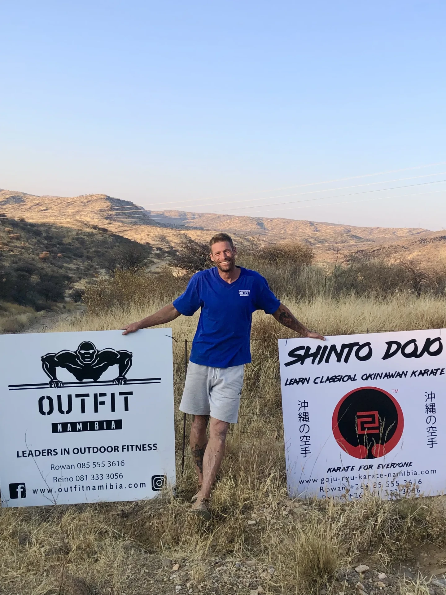 Rowan standing between the Outfit Namibia and Shinto Dojo signboards on the hillside