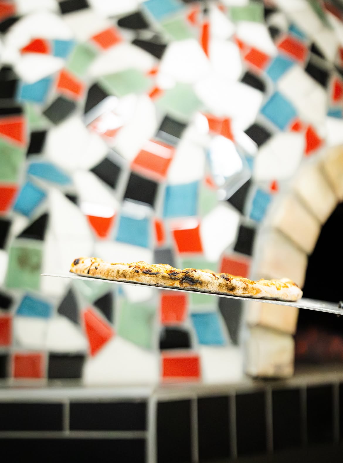 Pizza on a metal peel in front of the mosaic oven