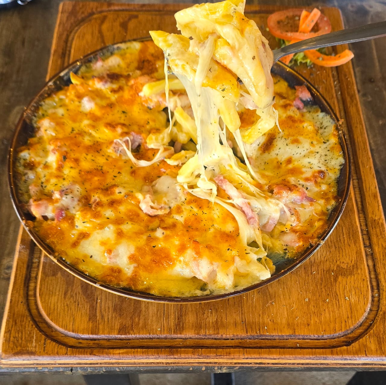 Baked plate loaded with melted cheese and bacon, lifted with a fork on a wooden serving board