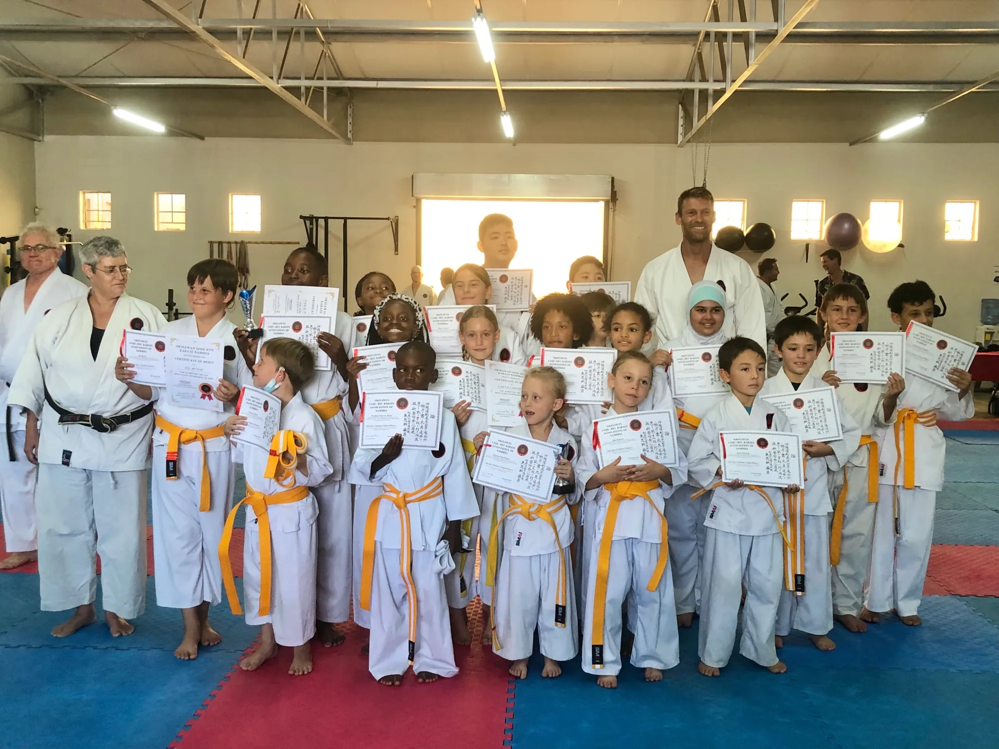 Children and instructors in white karate gi with yellow belts holding their grading certificates at Shinto Dojo